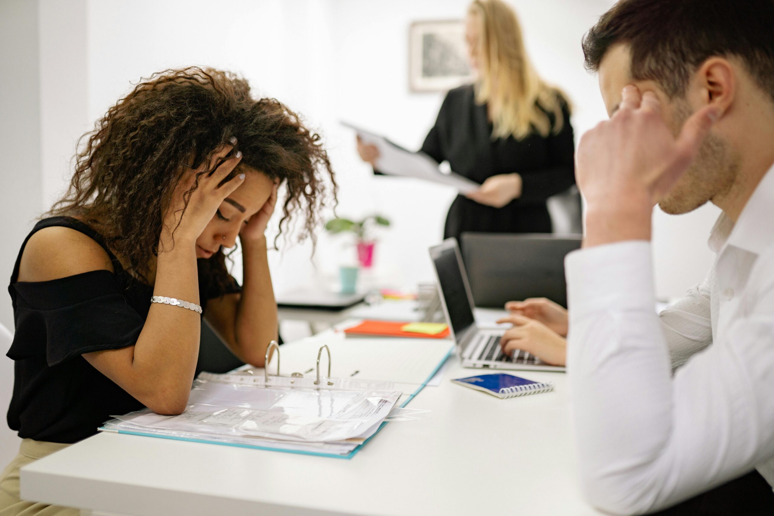 African American woman stressed at office desk with colleagues nearby, conveying workplace pressure.